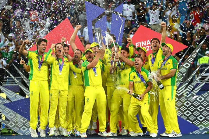 Players in yellow uniforms with green trim celebrating with champagne, flags and confetti. Spectators just visible behind.