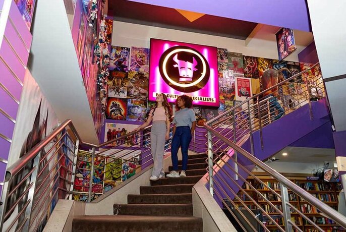 Two women walking down the stairs in a gaming shop with a pink neon sign.