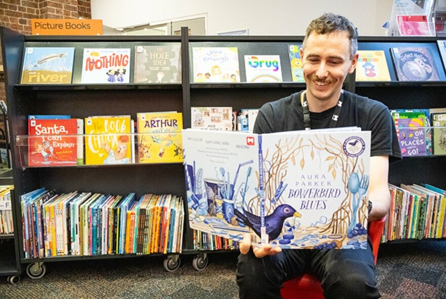 A smiling person holding up an illustrated book and reading from it in a library setting, shelves of books behind him.
