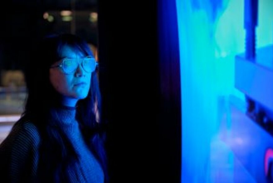 A woman wearing glasses stares blankly at a dark blue illuminated wall.