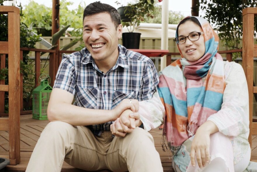 A smiling man, seated outdoors on a deck, holding the hand of a smiling woman, also seated, who wears a headscarf and glasses.