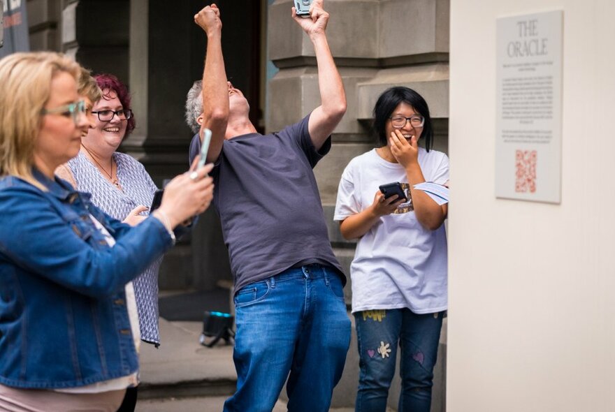 People stand around a wall with their phones out, one man leaning back and jubilantly raising his fists in the air. 