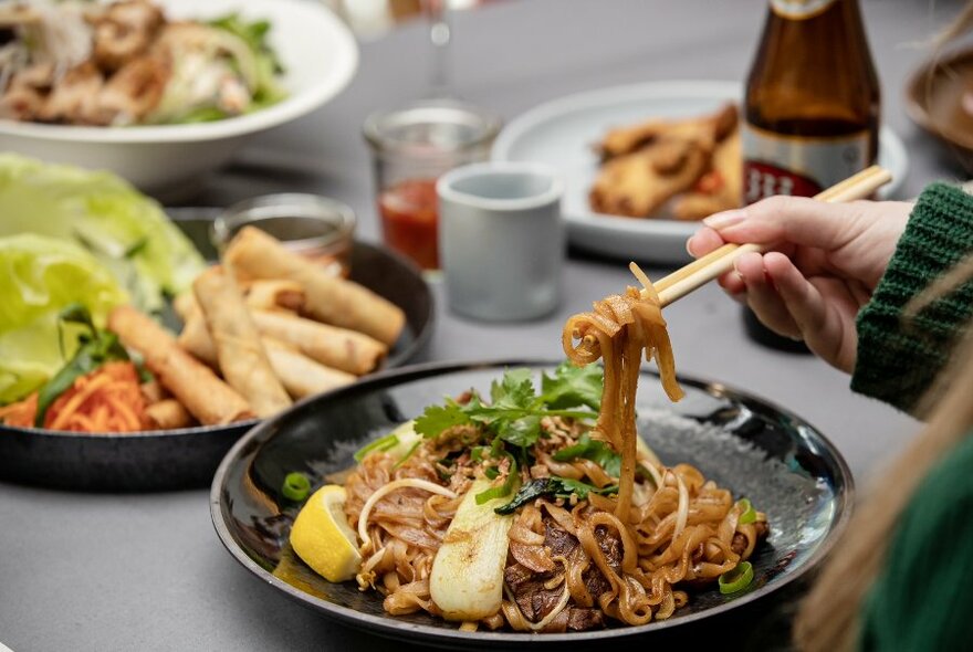 Someone lifting noodles with chopsticks at a table with multiple plates of Vietnamese food, beer and condiments. 