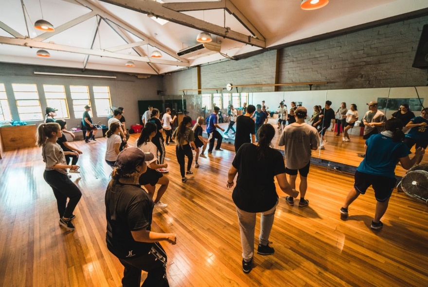 Group of people dancing on a wooden floor in a dance studio, all facing a full wall mirror.
