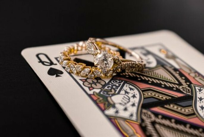 Two silver and gold rings encrusted with stones, resting an a Queen of Spades playing card.