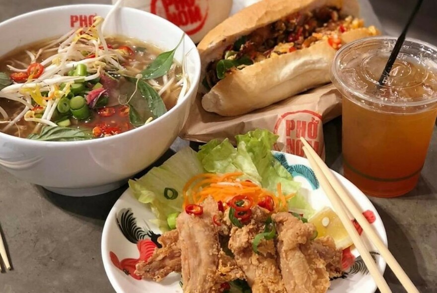 Bowl of pho, banh mi and chicken dish nestled with plastic cup of soft drink.
