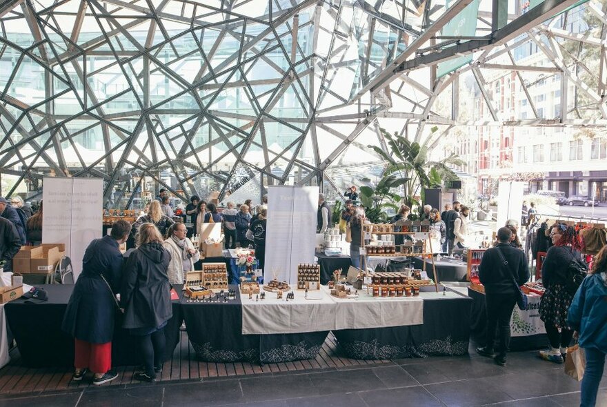 Market stalls and tables selling their assorted wares in the covered Atrium at Fed Square, with people browsing.