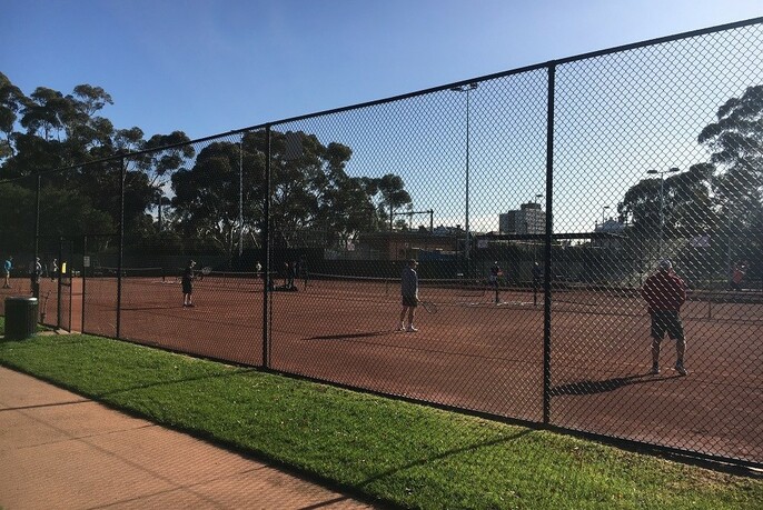 People playing tennis on red clay tennis courts behind a high fence.