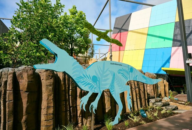 A blue cut out of a dinosaur in a garden outside the colourful Melbourne Museum building.