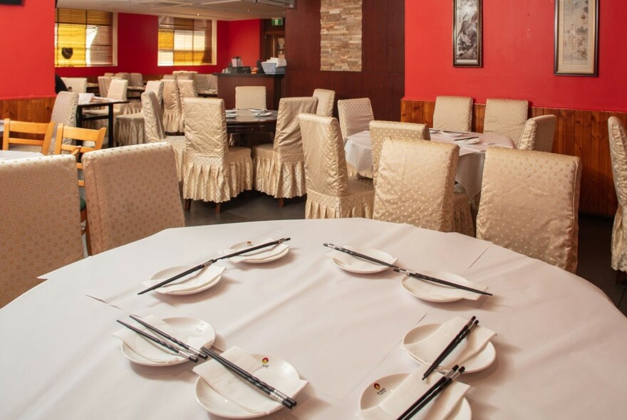 Plates and chopsticks on a round table with white cloth, chairs with white fabric covers in a room with red walls.