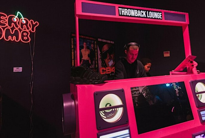 Pink and purple DJ booth, with man wearing headphones standing at centre. 