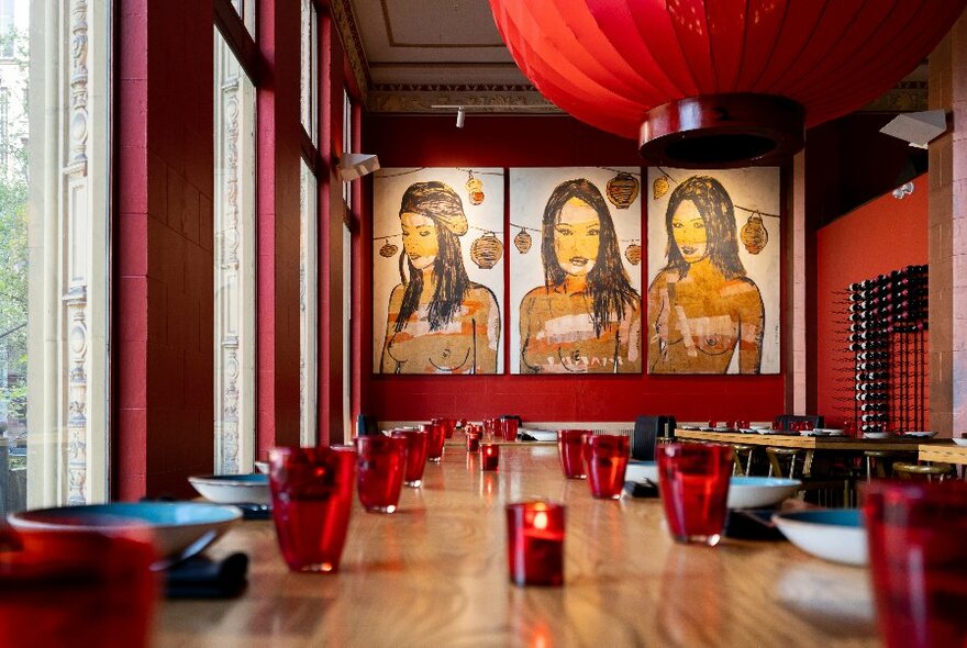 Interior of Red Spice Road with long wooden table and chairs, red ceiling lantern and three portraits at back of room.
