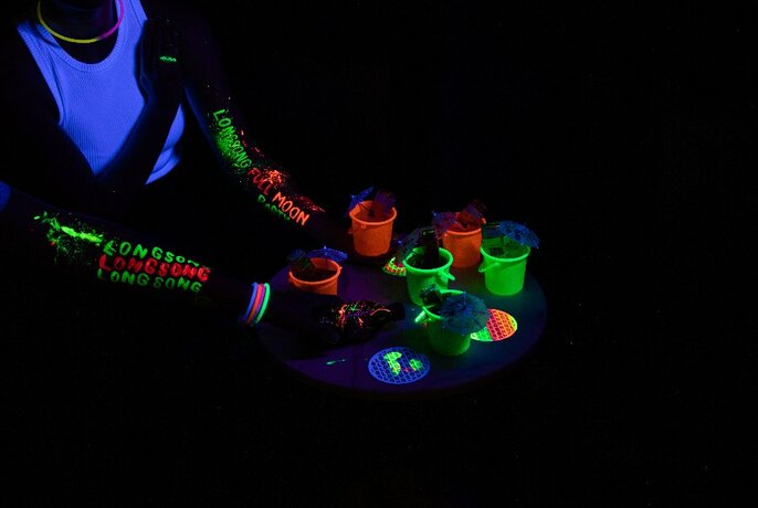 Torso and hands of person with UV-lit writing on arms and colourful cups on high table. 