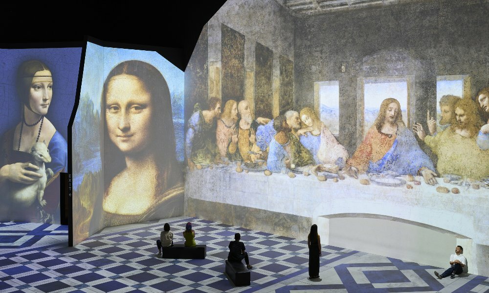 A group of people are in a digital projection art gallery looking at famous paintings including the Mona Lisa and The Last Supper