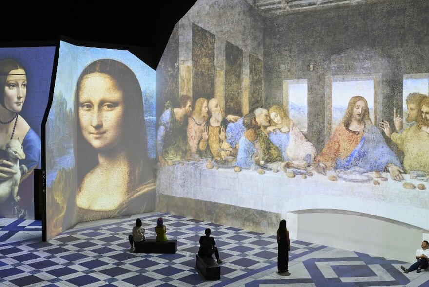 A group of people are in a digital projection art gallery looking at famous paintings including the Mona Lisa and The Last Supper