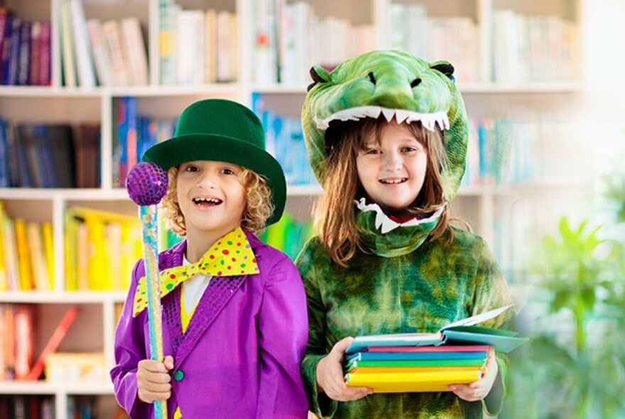 Two young smiling children dressed up us characters from Roald Dahl stories - Willy Wonka and a green crocodile - standing in a library setting with bookshelves behind them.