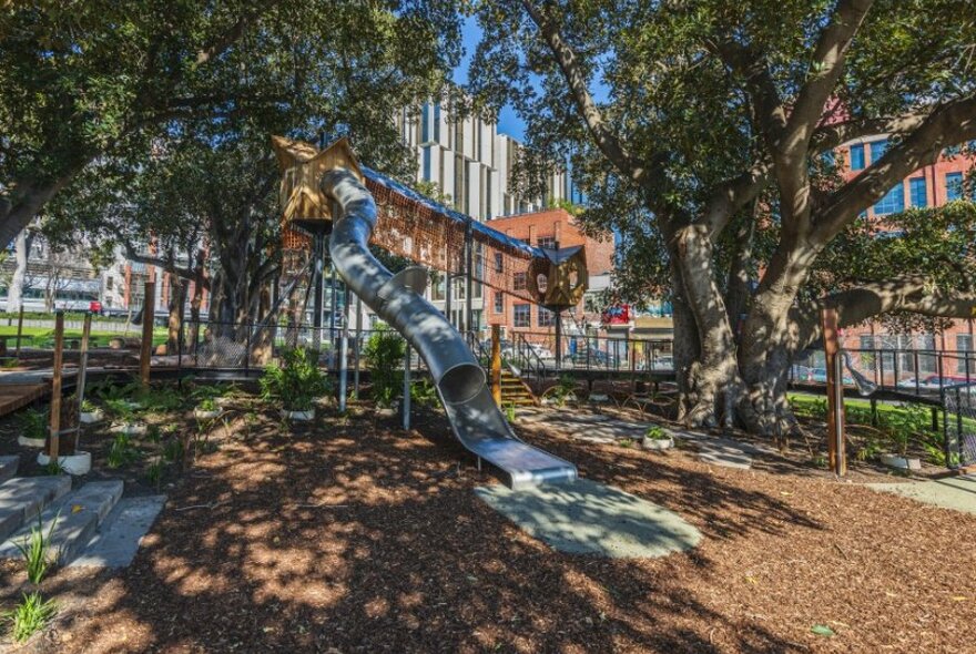 A shady playground with elevated walkway and a long slide.