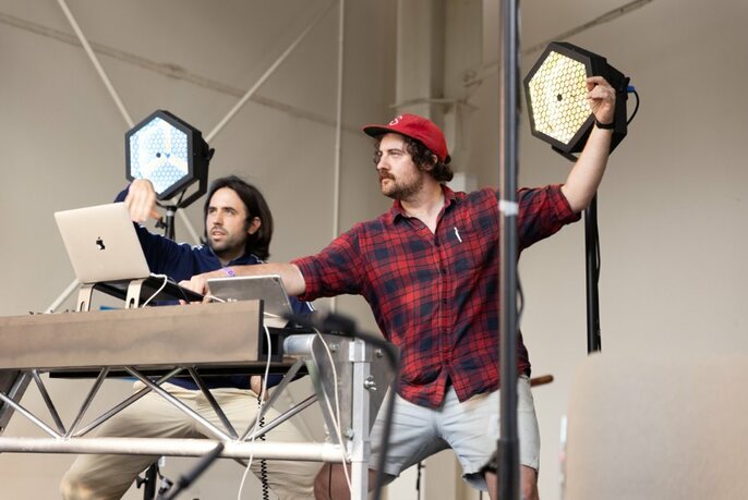 Two performers in casual clothes hold up hexagons while operating computers.