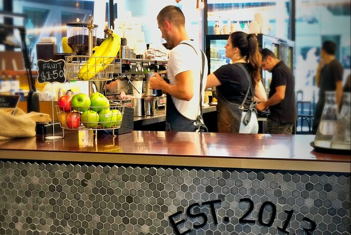 Barista making coffee using an espresso machine with a basket of apples and bananas on the counter. 