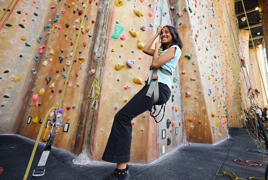 A smiling girl hanging from a harness on a rock climbing wall.
