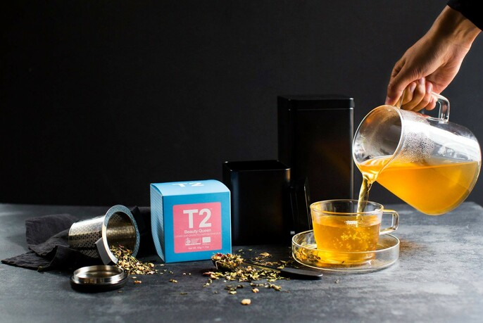Tea being poured into a glass teacup and a brightly-coloured packet of T2 tea.