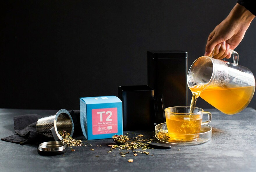 Tea being poured into a glass teacup and a brightly-coloured packet of T2 tea.