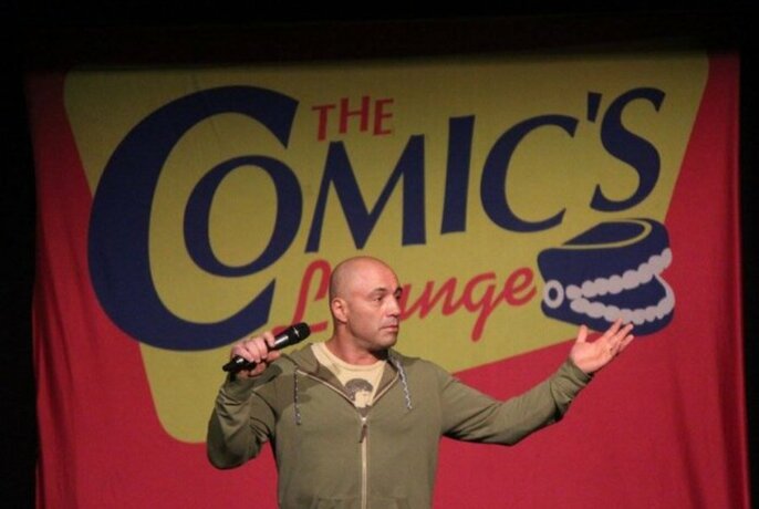 Comedian holding a microphone and performing on stage in front of a red, yellow and blue backdrop that features the words 'The Comic's Lounge' written on it.