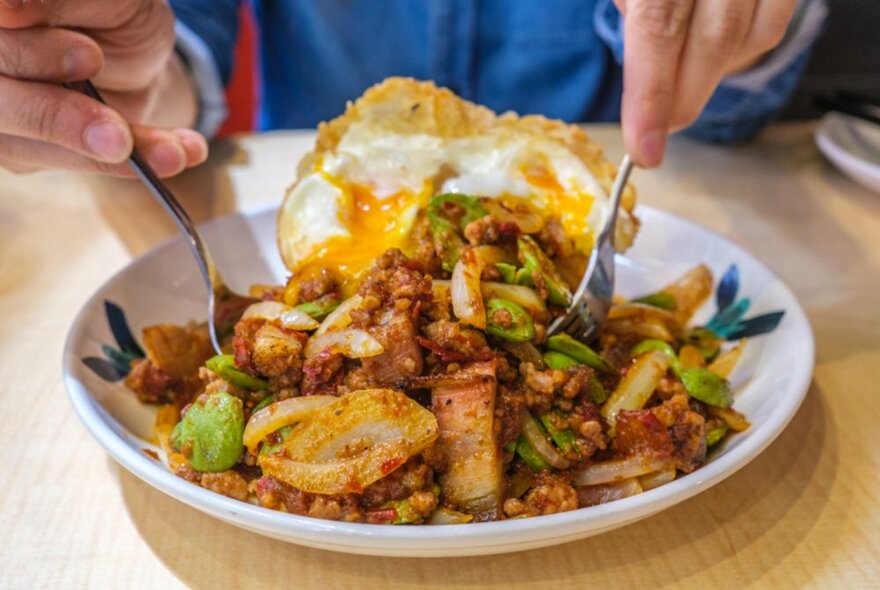 Close up shot of person sticking a knife and fork into a bowl of chicken and vegetables with a fried egg.