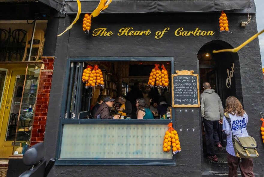 Cafe exterior with black paintwork and yellow lettering, bags of oranges strung from walls, people seated inside next to large open window as two customers enter through the arched doorway.