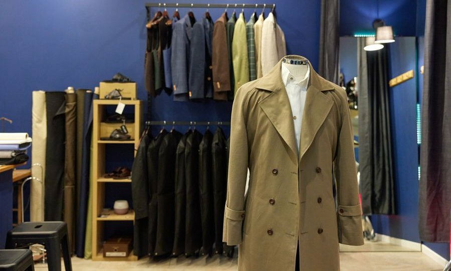 Wide shot of beige coat on mannequin, jackets and coats hanging in background against a blue wall.