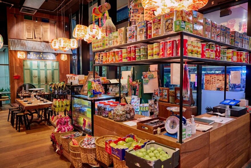 Interior of a colourful eatery showing shelves of food and produce for sale, like a mini-mart, with tables and chairs for dining up the back.