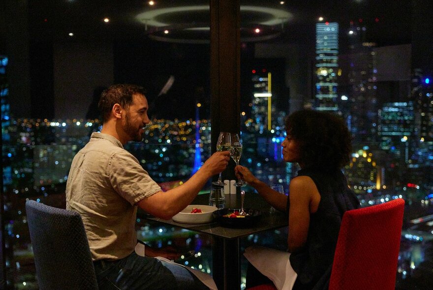 a couple are doing a cheers with champagne there is a city skyline as their view.