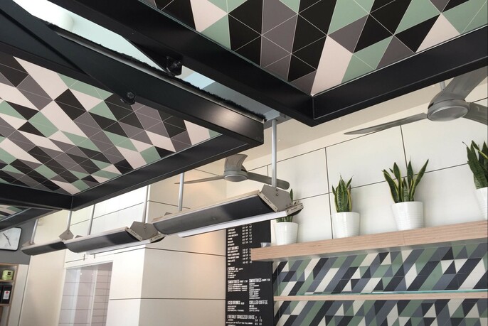 Black, green cream geometric patterned ceiling panels and wall tiles, with fans, green potted plants and black menu on wall.