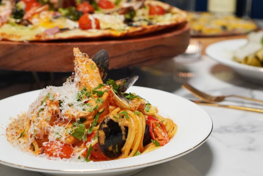 A bowl of pasta in foreground with a pizza in the background.