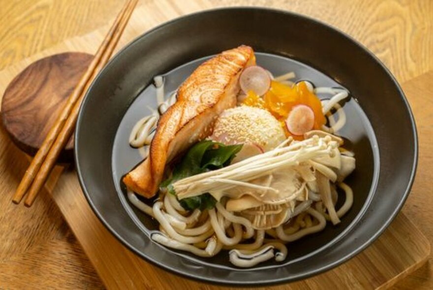 Black dish of fish and noodles with with chopsticks.