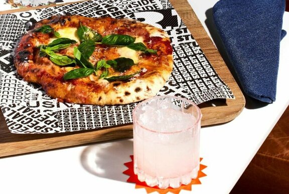 A margarita pizza on a wooden tray, topped with fresh basil, with a cocktail on a coaster in front of it.
