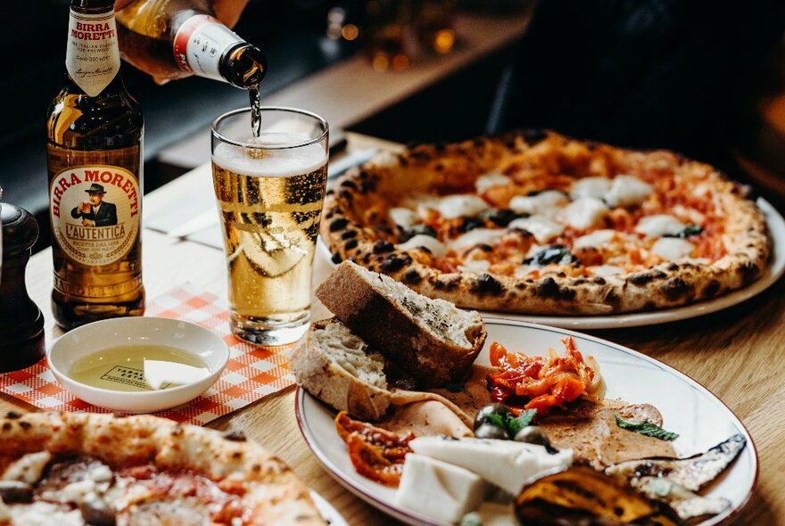 A table with several wood-fired pizzas and someone pouring a beer. 