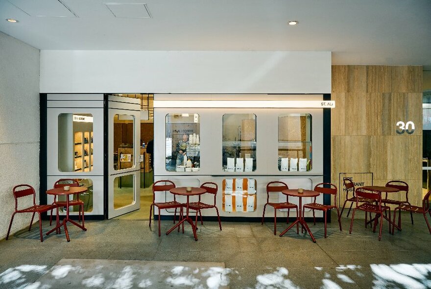 Exterior of cafe tucked into the corner area of a wall in a foyer space, with small red tables and chairs out the front.