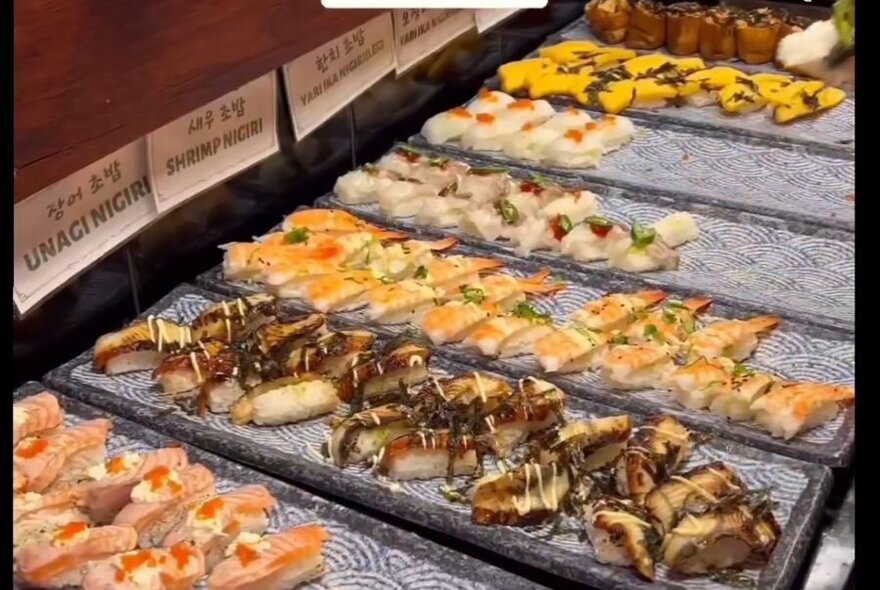 Platters of sushi lined up at a buffet display, with labels above them.