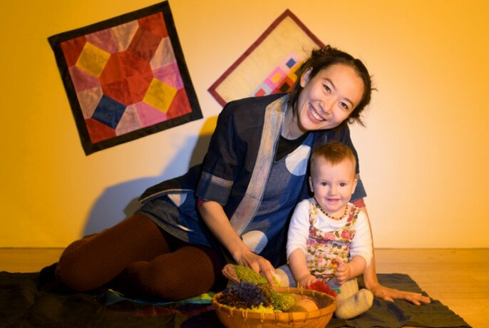 A small child and a grown-up sit on a mat with a bowl of fruit in a yellow room.