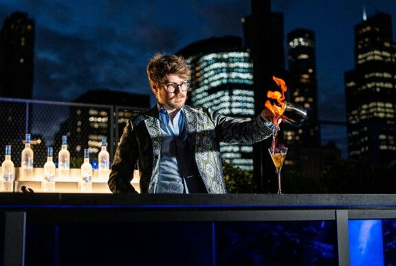 A bartender creating a flaming cocktail in a bar with lit up vodka bottles and city lights in the background.