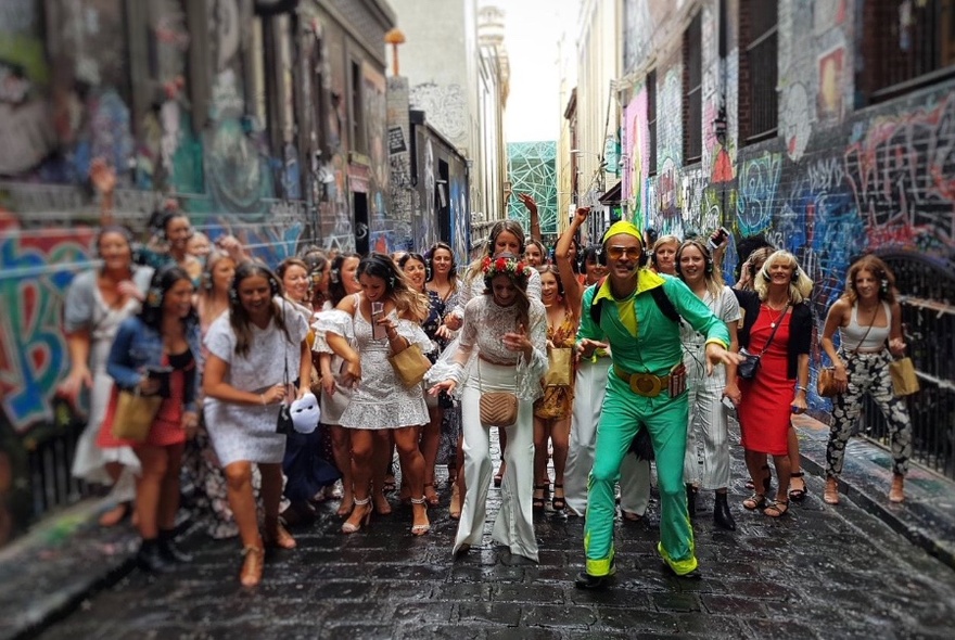 Group dancing in a laneway, led by man in bright green and yellow shiny outfit.