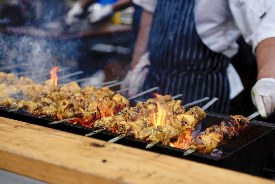 A chef cooking meat skewers over a flaming grill. 