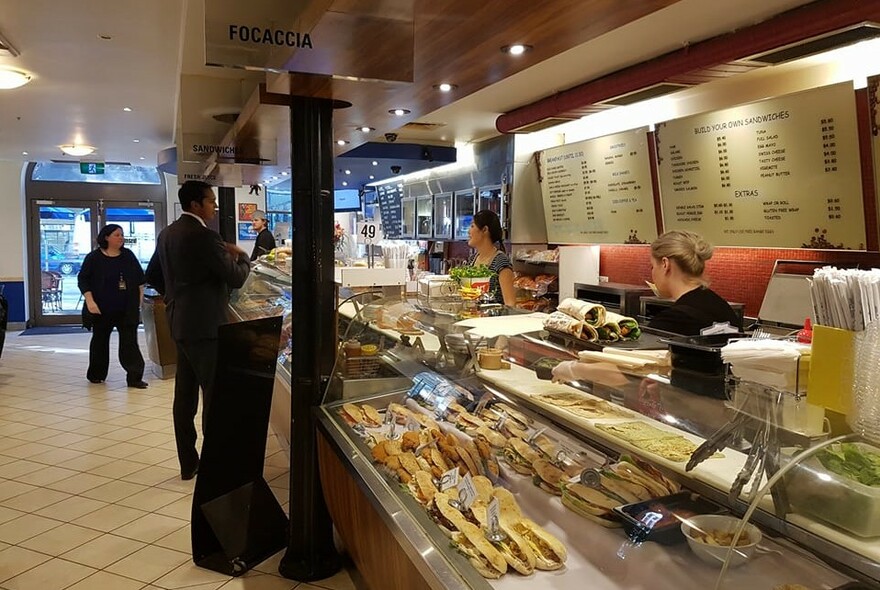Customers ordering at a cafe food preparation counter topped with sandwiches and wraps.
