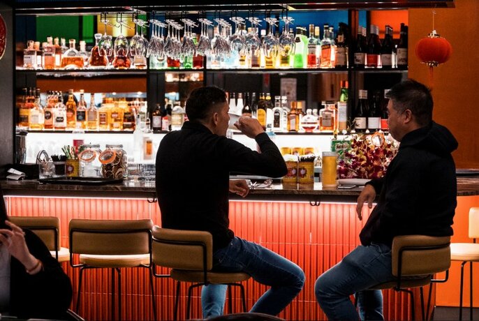 Two people sitting on high stools and drinking at a bar, a wall of bottles behind the bar area.