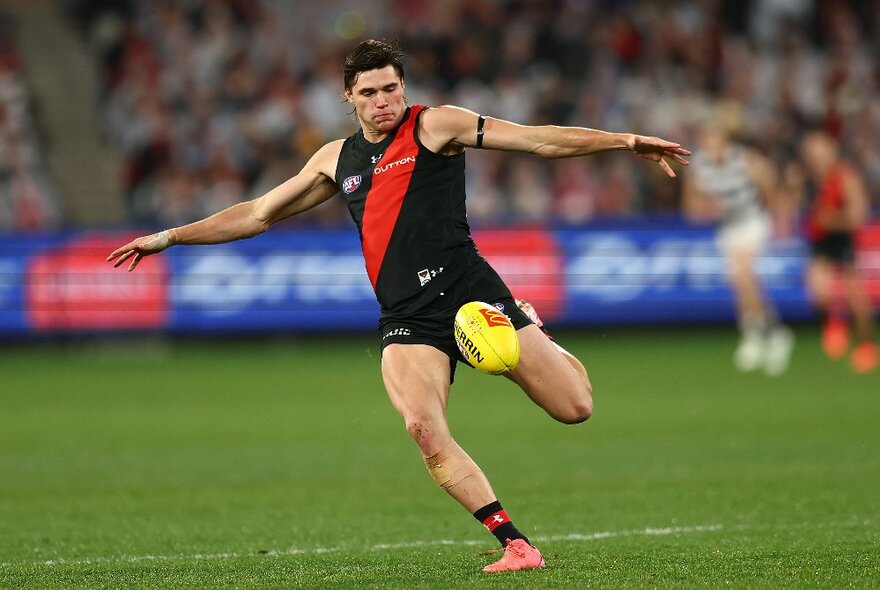 Essendon AFL football player wearing red and black, kicking a yellow ball during a match.