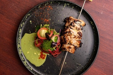 Black plate of food with skewered meat, salad and green sauce.