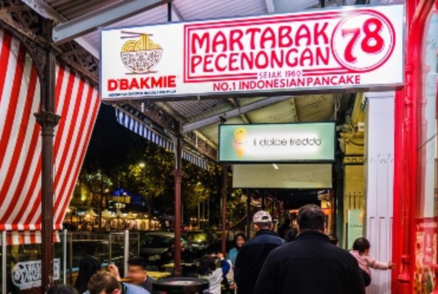 The exterior of Martabak Pecenongan 78 in Lygon Street Carlton, with illuminated shop signage hanging from the awning, people dining outside on tables and people walking past; night scene.