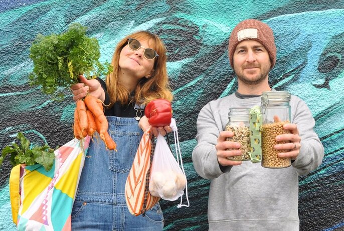 Two people standing against bluey-green painted wall, one on left holding up veggies, one on right holding glass jars of produce.