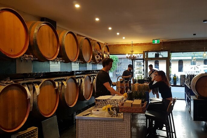 Interior of shop showing rows of large wooden wine barrels behind the shop counter with two staff members, and a shop patron sitting on a stool.